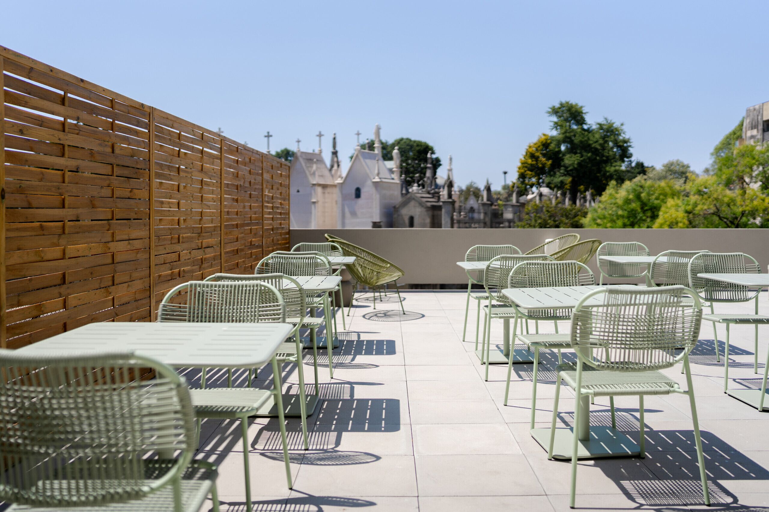 A rooftop patio with metal tables and chairs, a brick wall, and a view of a cemetery with various buildings and trees.
