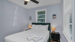 Un lit bien fait, avec du linge de lit blanc et deux serviettes enroulées, une tête de lit recouverte d’un tissu vert, deux lampes de chevet, une fenêtre avec des stores et un ventilateur de plafond.