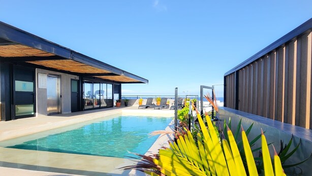 Un área de piscina moderna con vistas despejadas al mar, una zona de estar cubierta y un edificio con amplios ventanales.