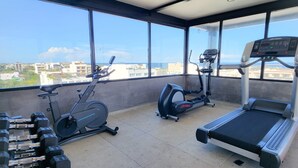 Un gimnasio con equipo de ejercicio, incluyendo mancuernas, una máquina elíptica y una cinta de correr, con grandes ventanales que ofrecen vista a la ciudad.