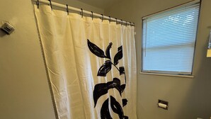 Une salle de bain avec un rideau de douche blanc orné d’un motif de feuilles noires, des toilettes et une fenêtre avec des stores.