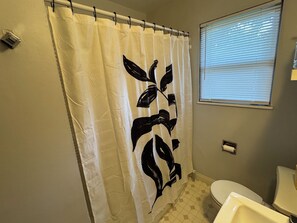 Een badkamer met een witte douchegordijn met een zwart bladmotief, een toilet en een raam met zonwering.