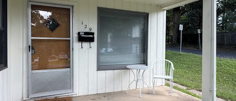 A house with a front door, a mailbox, a window with blinds, a small table and chair, and a welcome mat.
