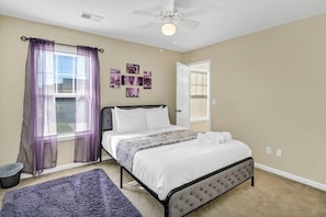 Une chambre à coucher avec un lit, un ventilateur de plafond, une fenêtre avec des rideaux violets et un mur orné de tableaux.