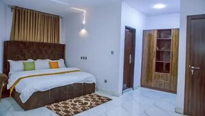 A bedroom with a large bed, a headboard, a bedside table, a nightstand, a patterned rug, and a wardrobe with a mirror. - TAJ SUITES  (Ado Ekiti)