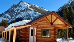 A wooden cabin with a pitched roof, a door, and a window, surrounded by snow and mountains. - Index Peak Cabin (Silver Gate)