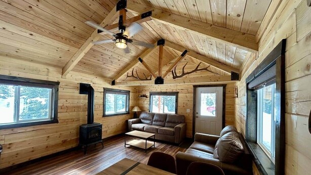 A wooden cabin interior with a high ceiling, a fireplace, a sofa, and a dining table. - Index Peak Cabin (Silver Gate)