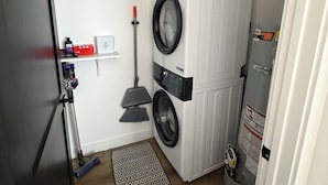 Une buanderie équipée d’un lave-linge et d’un sèche-linge superposés, d’un balai, d’une serpillière et d’une petite étagère contenant des produits de nettoyage.