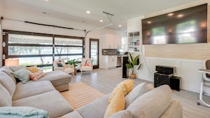 A modern living room with a large sectional sofa, a flat-screen TV mounted on the wall, and a kitchen in the background. - Waterfront Deck w/ Fire Pit: Lake Spokane Gem! (Nine Mile Falls)