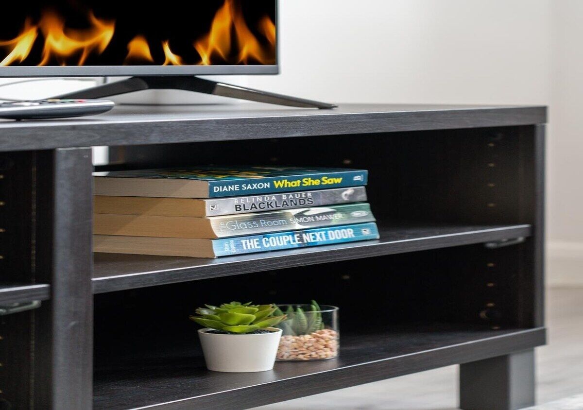 A black TV stand with a flat-screen TV displaying flames. On the stand, there are books titled 'What She Saw', 'Black Hills', 'Glass Room', and 'The Couple Next Door'.