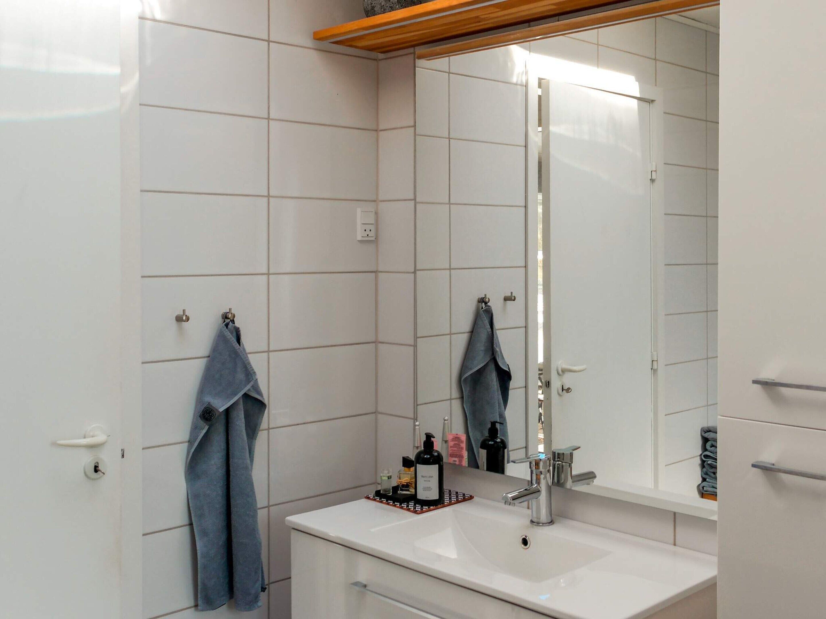 Ein Badezimmer mit weißen Fliesen, einem Waschbecken mit Spiegel darüber und zwei Handtüchern, die an Haken hängen.