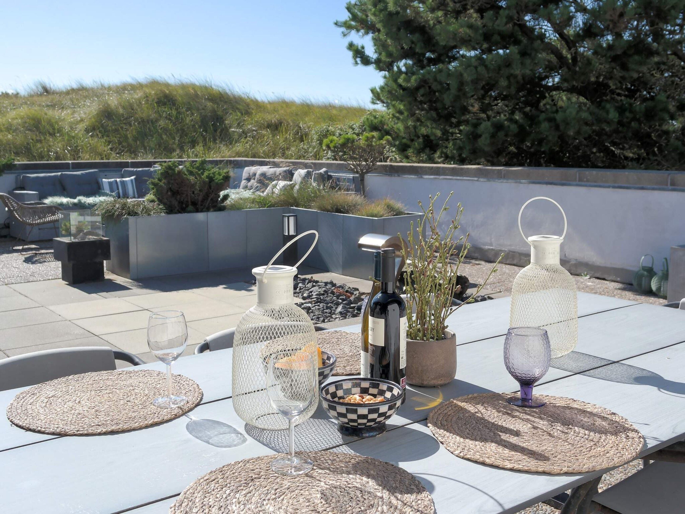 En udendørs terrasse med et spisebord dækket med vævede underlag, drikkeglas og dekorative genstande.