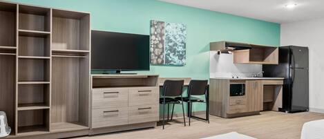 Une chambre d’hôtel moderne avec un mur d’accent de couleur turquoise, une armoire en bois, un bureau avec deux chaises, un réfrigérateur noir et un téléviseur à écran plat.