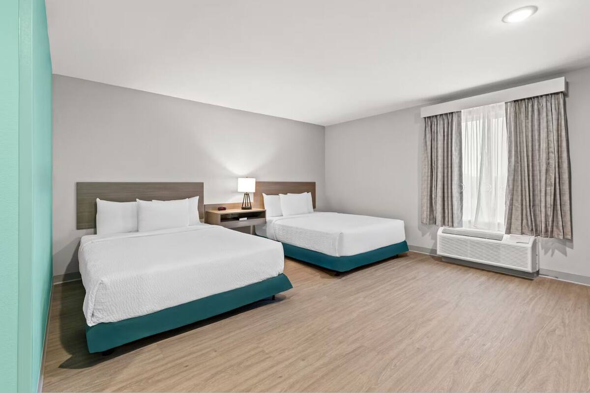 Habitación de hotel con dos camas, una mesita de noche con lámpara, una ventana con cortinas y un radiador.