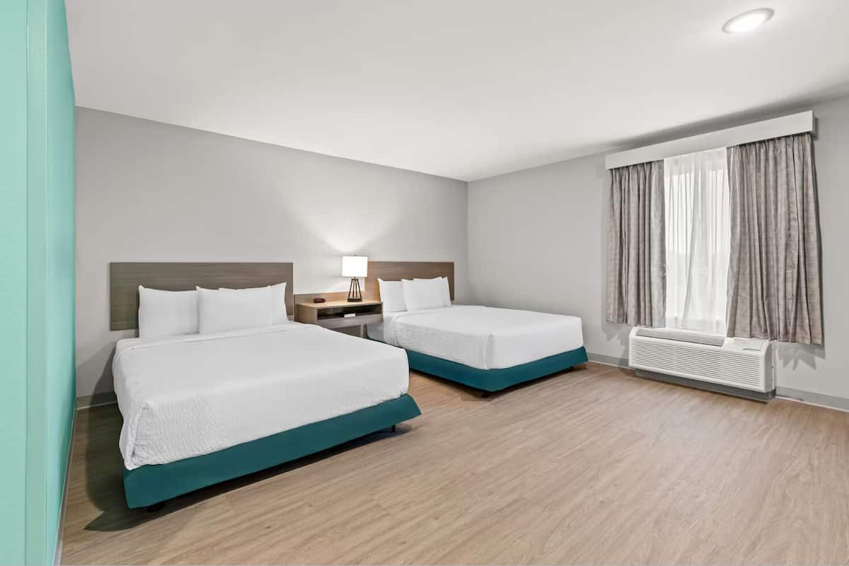 A hotel room with two beds, a nightstand with a lamp, a window with curtains, and a radiator.