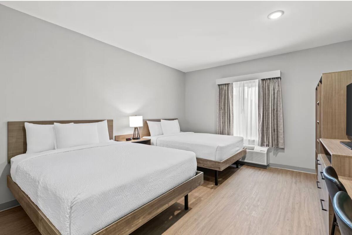 Habitación de hotel con dos camas, un escritorio, una silla, una lámpara y una ventana con cortinas.