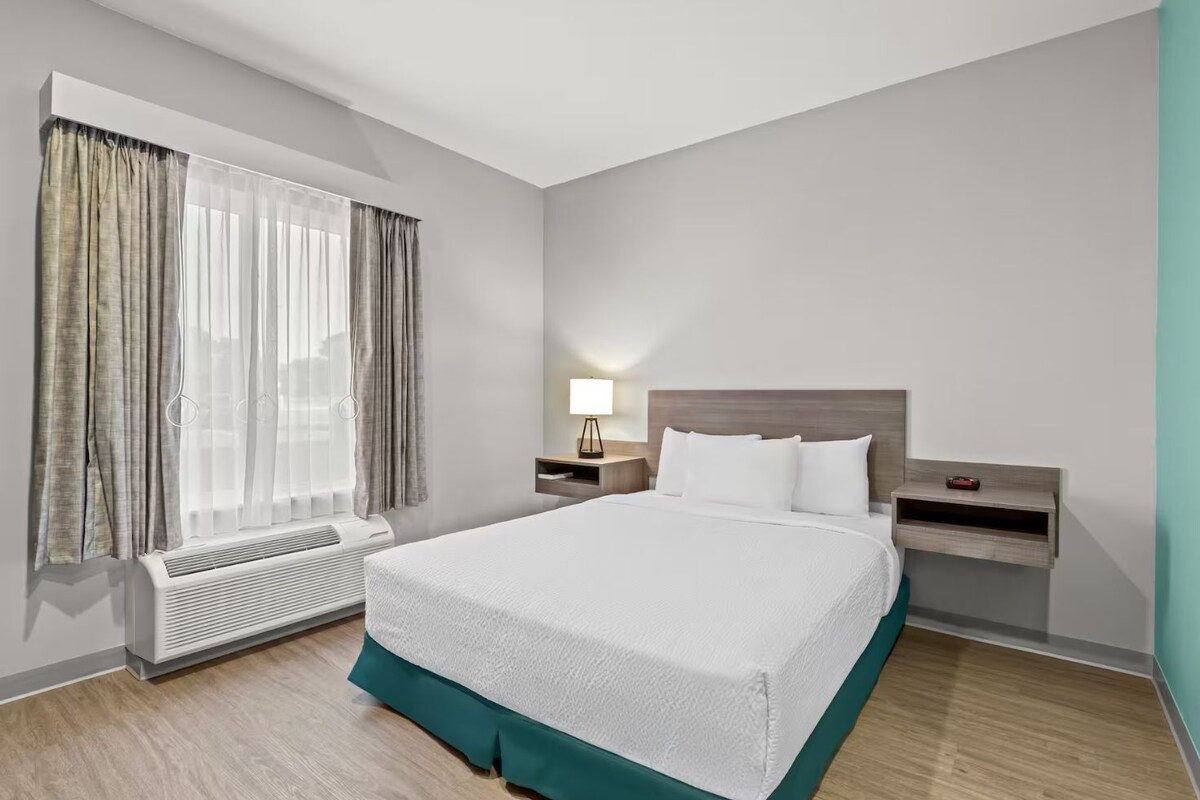 A modern hotel room with a large bed, a bedside table with a lamp, a heater, and a window with curtains.
