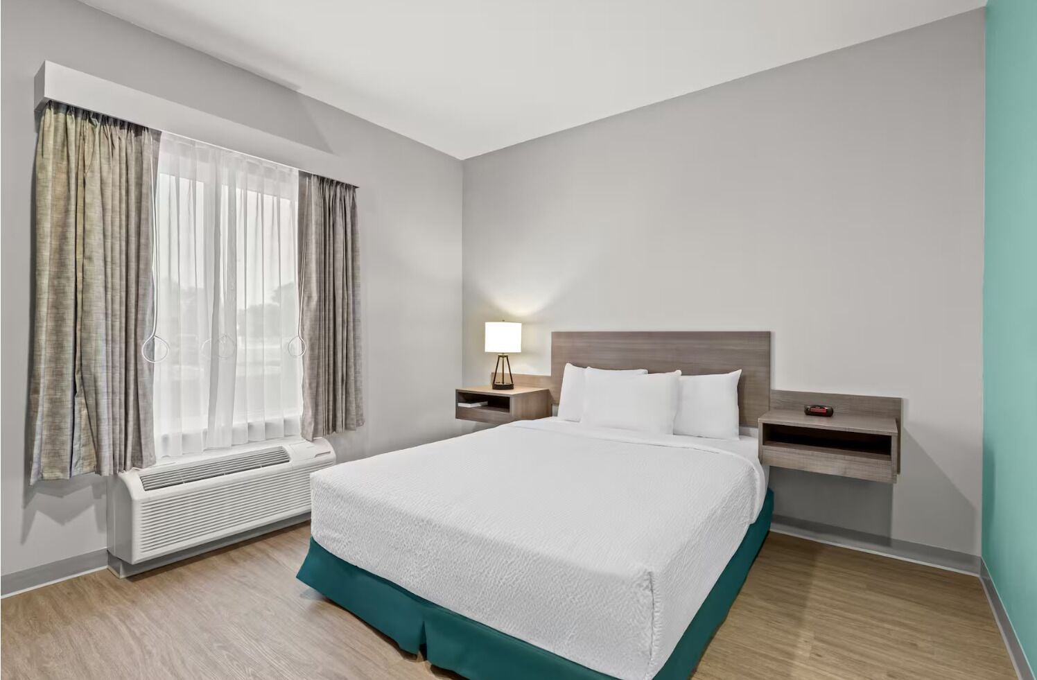 Una habitación de hotel moderna con una cama grande, una mesita de noche con lámpara, un calentador y una ventana con cortinas.