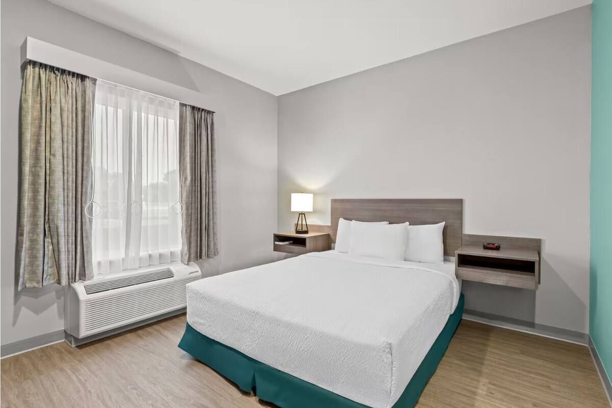 Una habitación de hotel moderna con una cama grande, una mesita de noche con lámpara, un calentador y una ventana con cortinas.