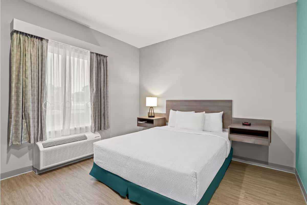 A modern hotel room with a large bed, a bedside table with a lamp, a heater, and a window with curtains.