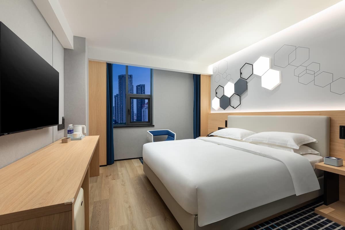 A modern hotel room with a large bed, a desk, a flat-screen TV, and a city view.