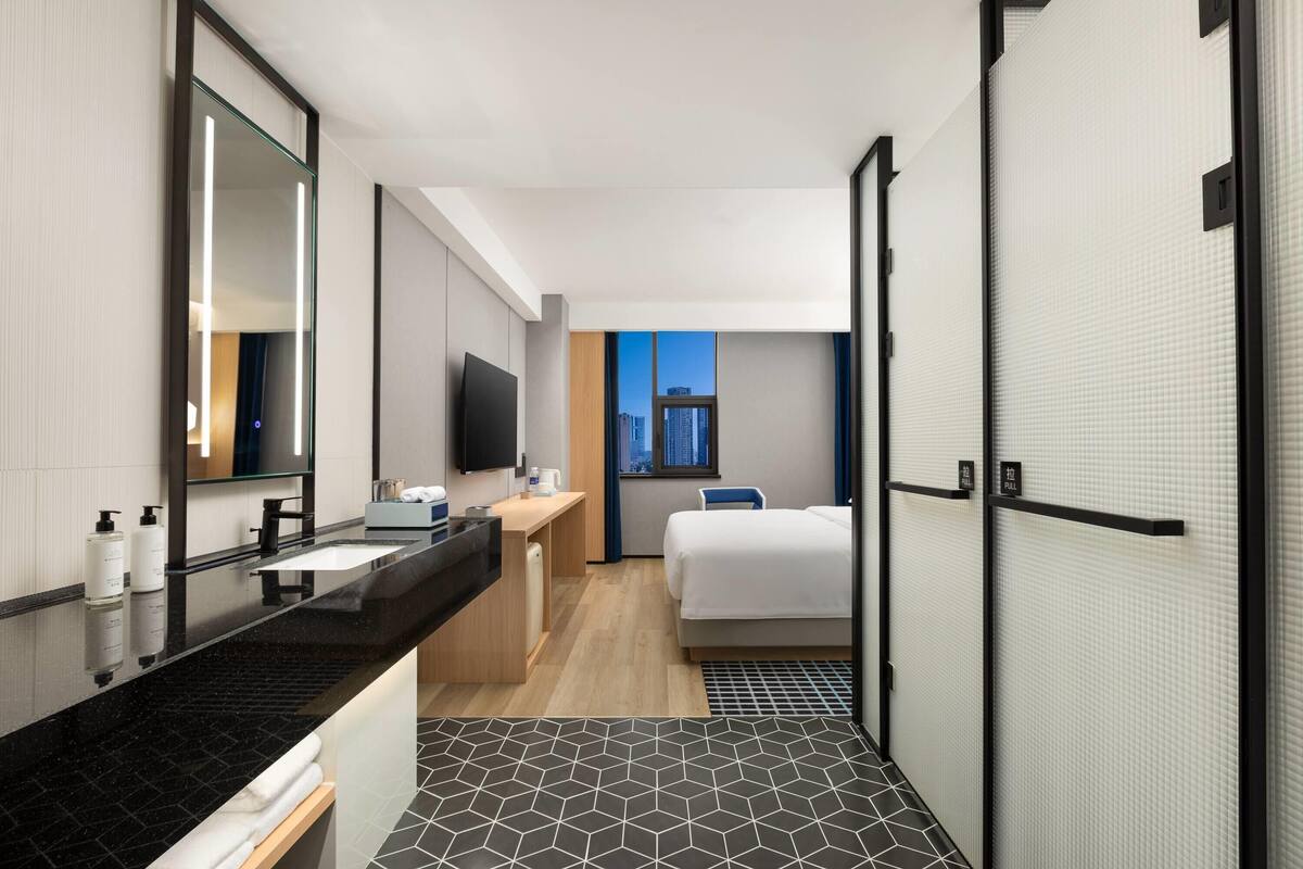 A modern hotel room with a large mirror, a black marble vanity, a bed, and a view of the cityscape through a window.