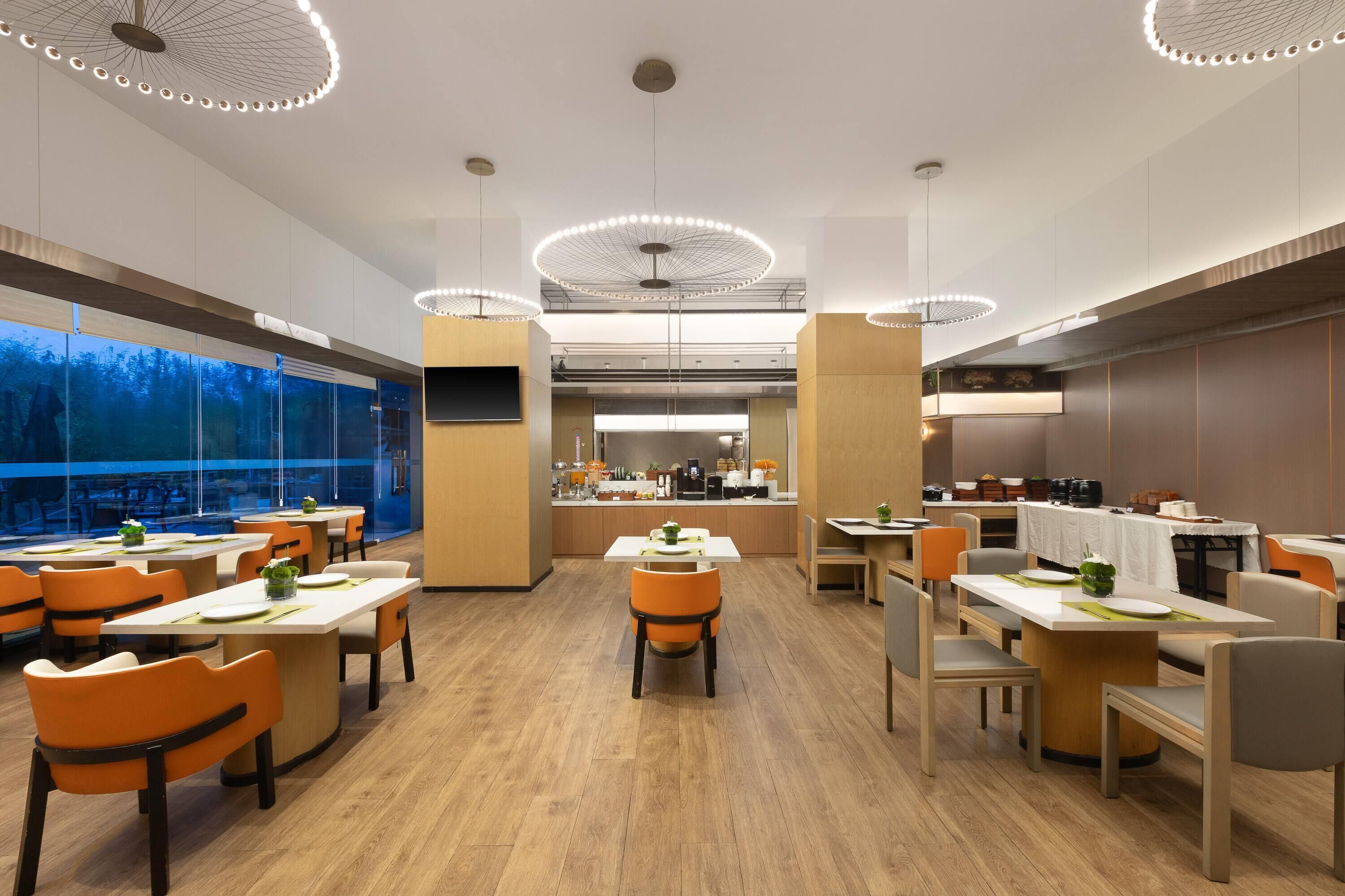 A modern dining area with multiple tables and chairs, a kitchen in the background, and large windows offering a view.