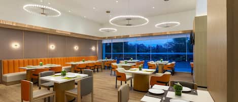 Uma área de refeições moderna, com mesas redondas, assentos em cabines laranja e iluminação pendente.