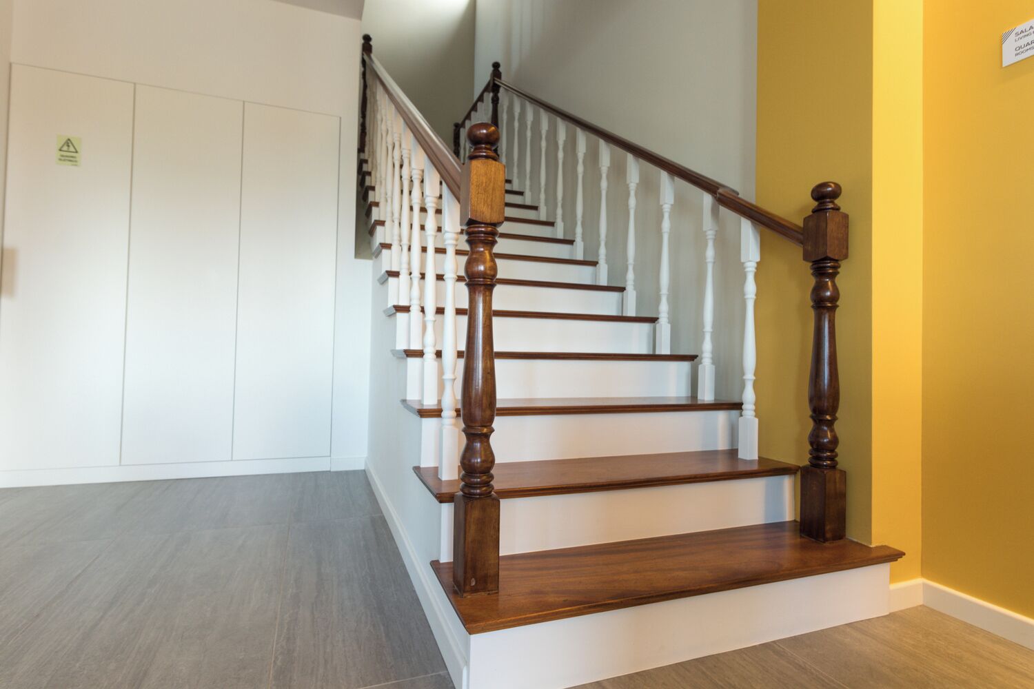Una escalera de madera con balaustres blancos y barandilla de madera, que conduce a un rellano con pared blanca y una pared de acento amarilla.