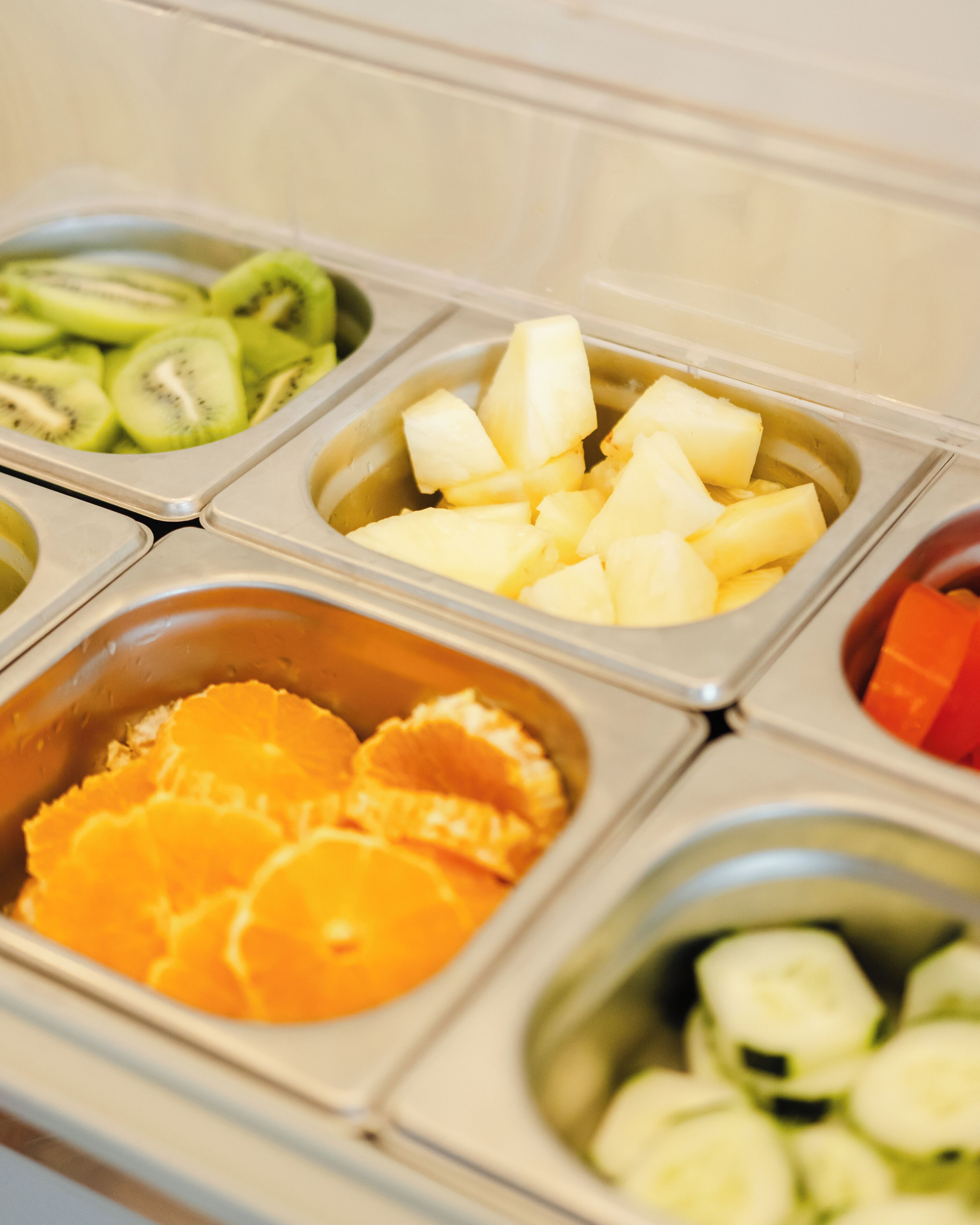 En buffet med frugt og grønt, inklusive kiwi, ananas, orangeskiver og agurk, serveret i separate beholdere.