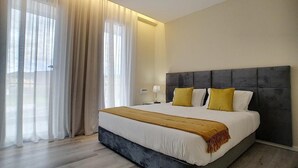 Une chambre d’hôtel moderne avec un grand lit, une tête de lit rembourrée de couleur grise et une vue sur l’extérieur à travers des rideaux transparents.