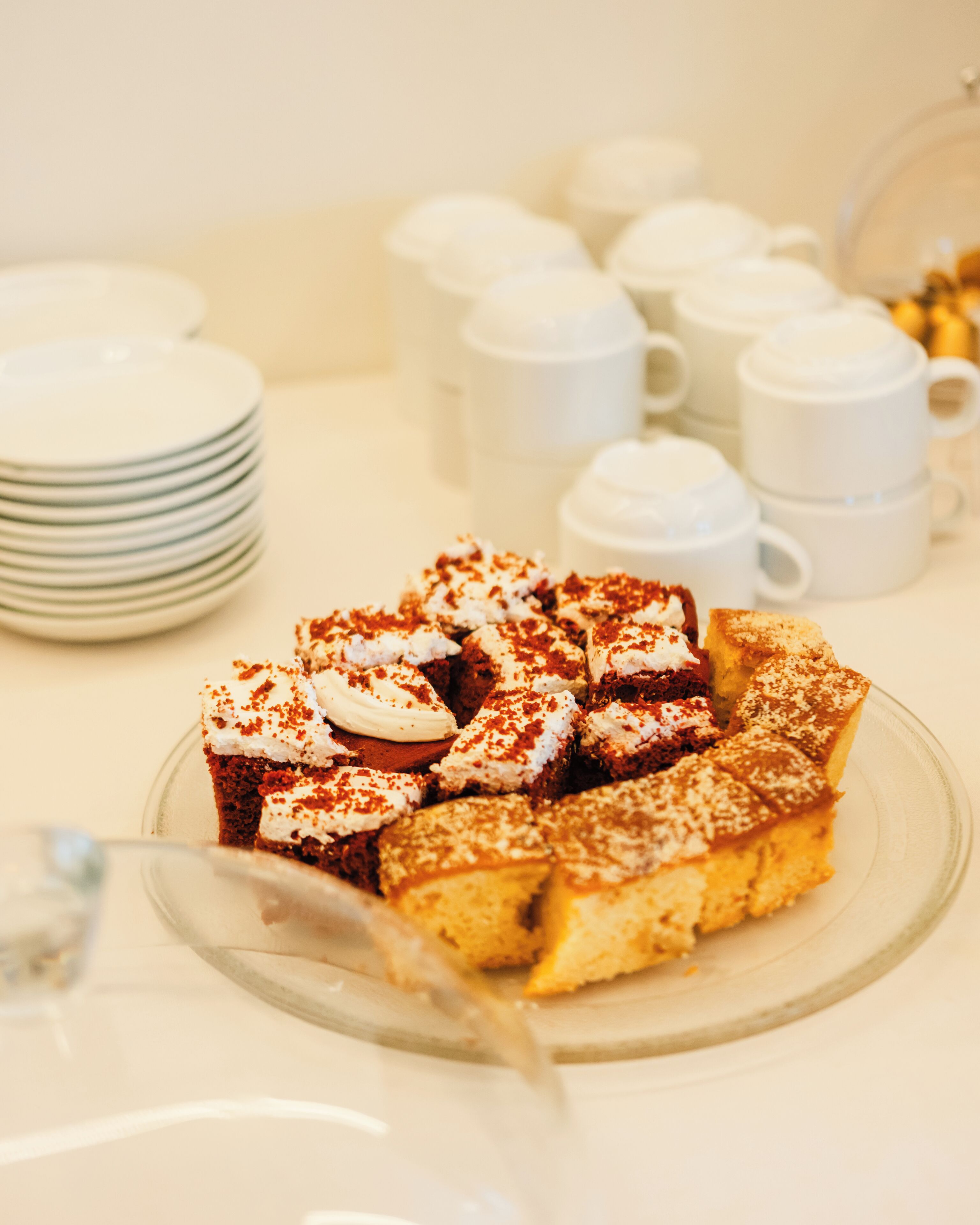 Eine Dessertplatte mit verschiedenen Kuchen und Gebäck, eine Stapel Teller sowie ein Satz weißer Tassen und Untertassen.