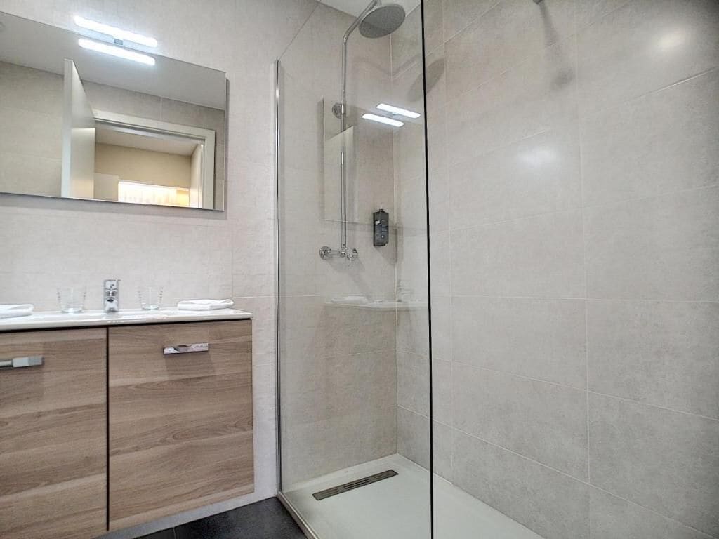 Ein modernes Badezimmer mit einer Glaskabine zur Dusche, einem hölzernen Waschtisch mit Spiegel und einem Fliesenboden.