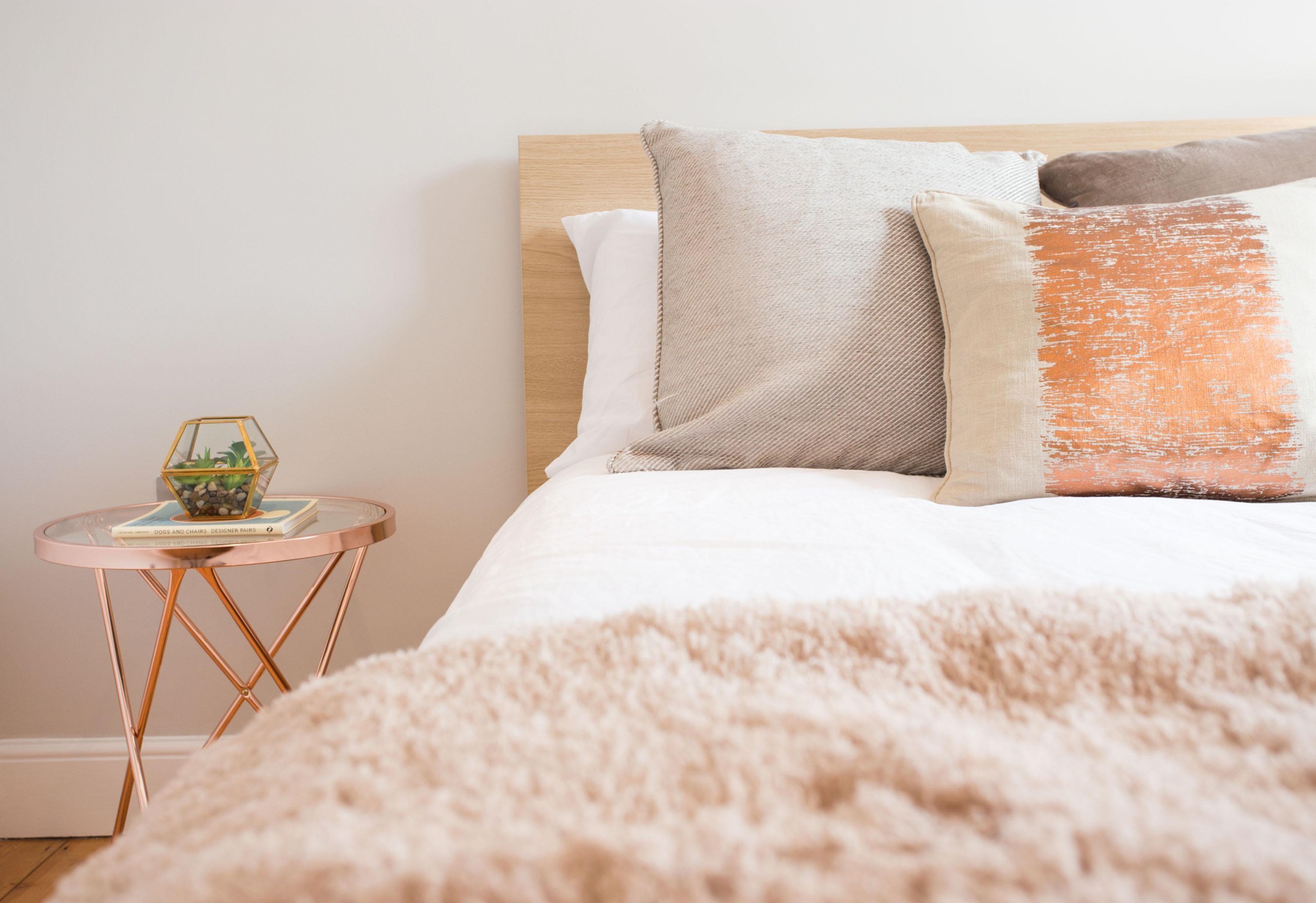 A neatly made bed with a fluffy beige blanket, a wooden headboard, and two pillows. There is a small round table with a glass top and a book beside the bed.