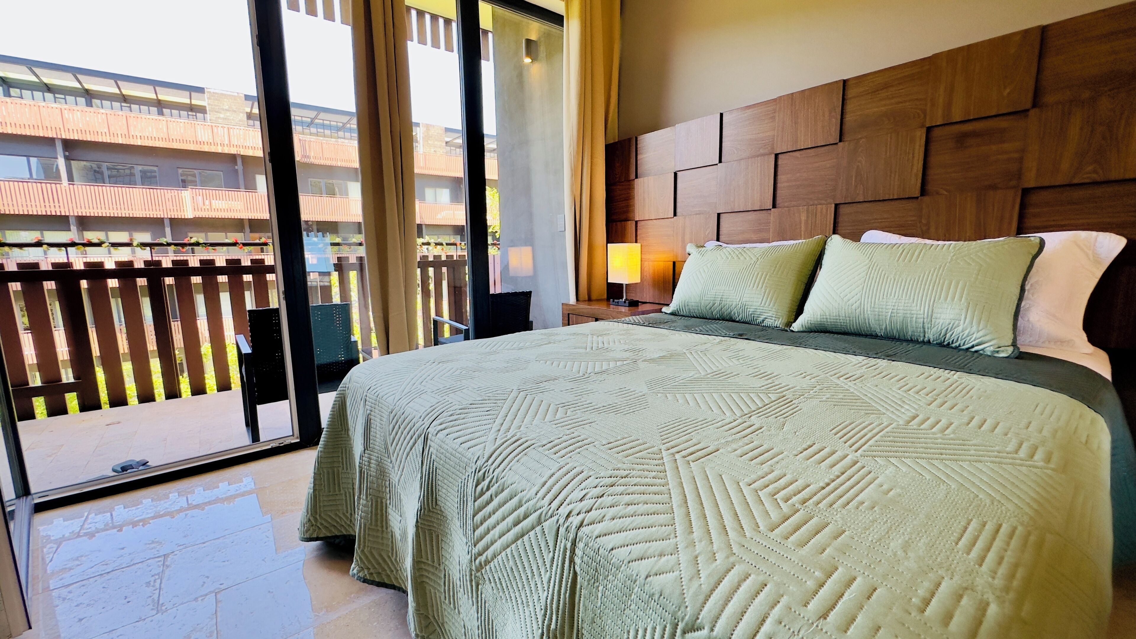 Een hotelkamer met een groot bed, een houten hoofdbord, een nachtkastje met een lamp en uitzicht op een balkon met een leuning.