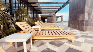Een rij houten lounge stoelen en tafels onder een pergola, met een geruite vloer en uitzicht op de zee.