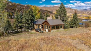 Une cabane dotée d’une cheminée en pierre, de grandes fenêtres et d’une terrasse aménagée avec du mobilier d’extérieur.
