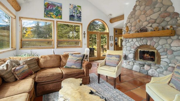 Un salon chaleureux avec une cheminée en pierre, des meubles en osier et de grandes fenêtres donnant sur l’extérieur.