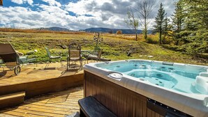Un bain à remous sur une terrasse en bois, agrémentée de mobilier d’extérieur, le tout entouré d’un paysage naturel avec des arbres et des montagnes en arrière-plan.