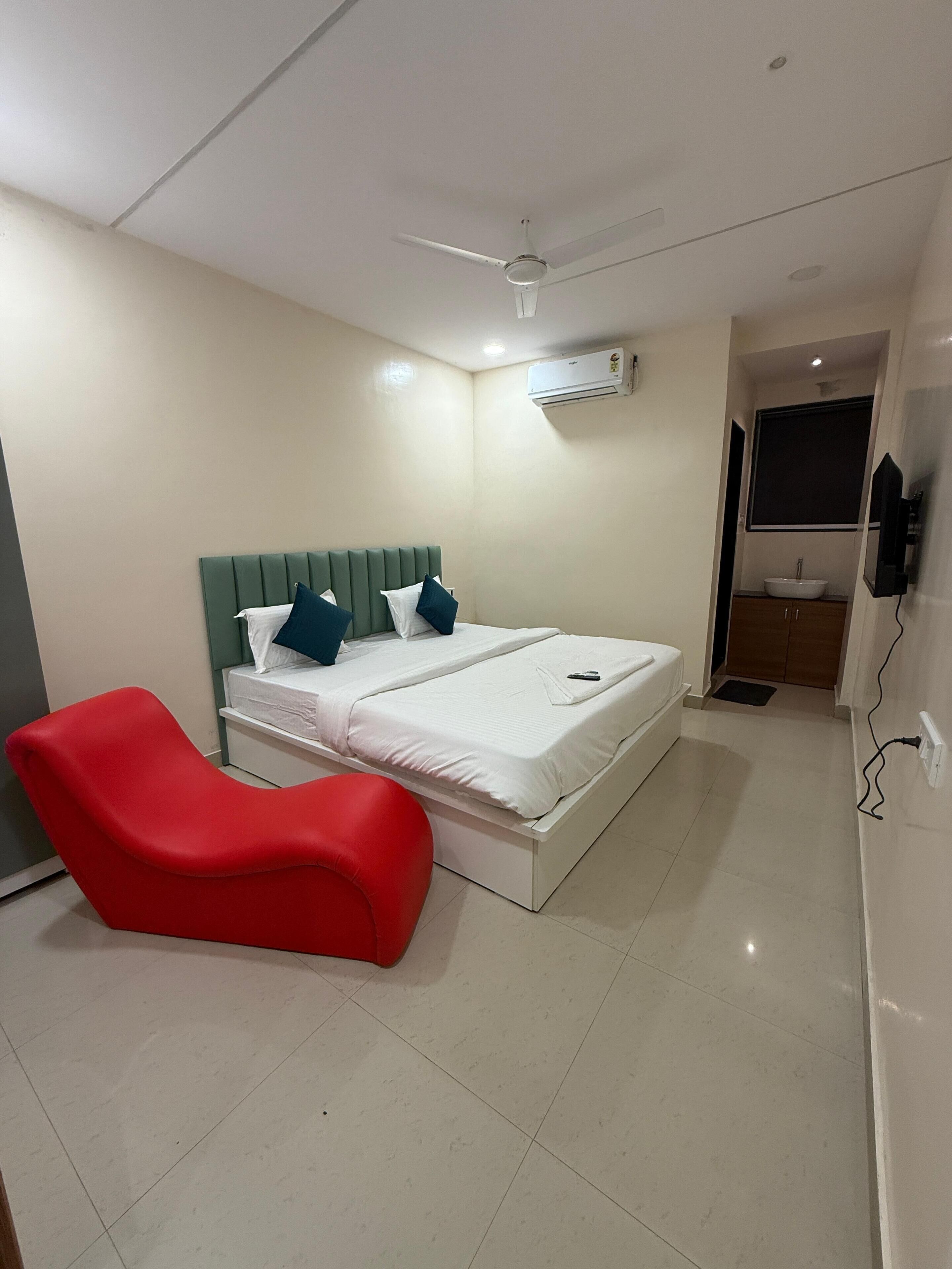 Un dormitorio con una cama, una silla roja, un ventilador de techo, un equipo de aire acondicionado y un televisor.