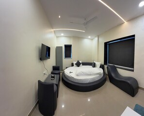 A modern bedroom with a circular bed, a flat-screen TV, a ceiling fan, and a large window. - Hotel Elite Stay - Mahalunge Pune  (Paud)