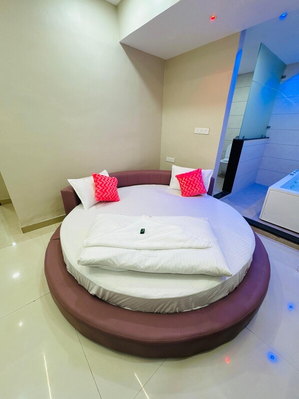 A circular bed with a brown headboard and white bedding, a small bedside table, and a view of a bathroom with a bathtub.