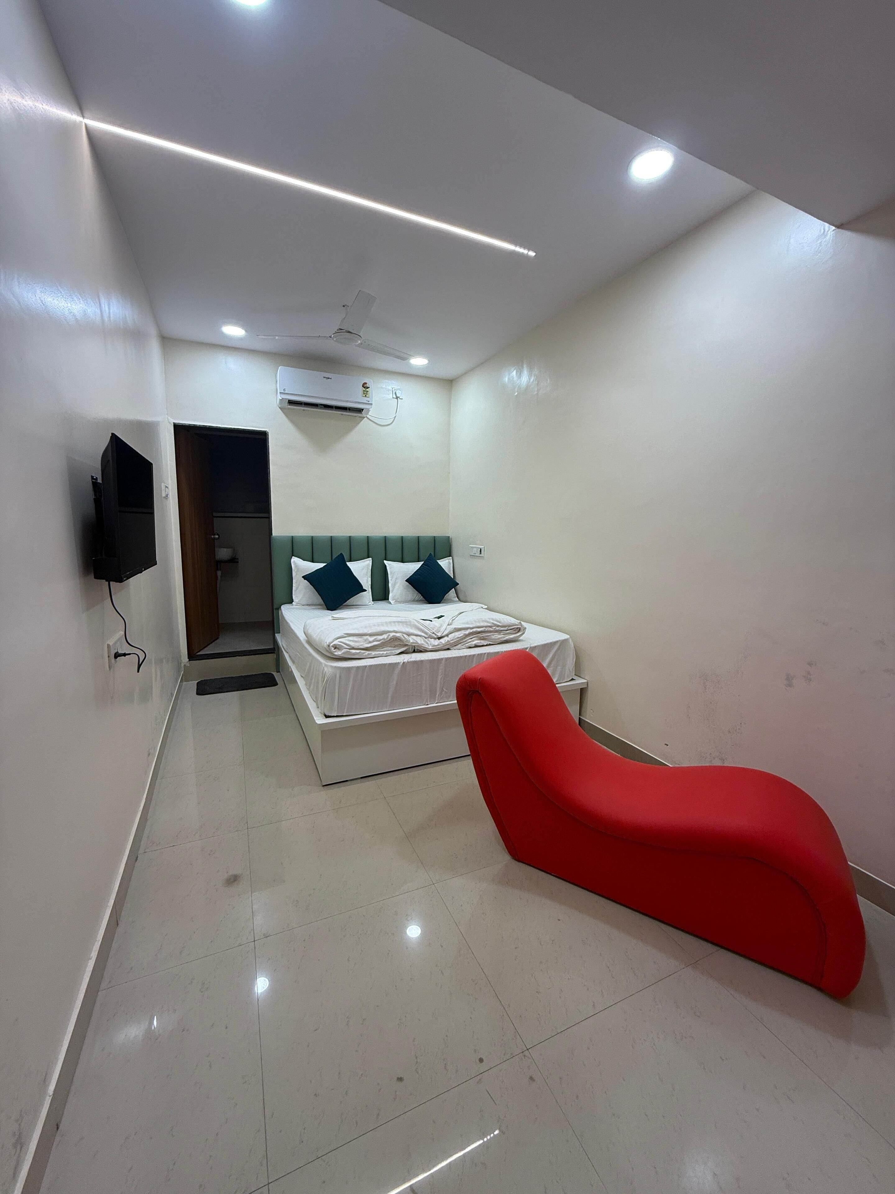 Una moderna camera d'albergo con un letto, una poltrona rossa, una TV a schermo piatto e un condizionatore d'aria.