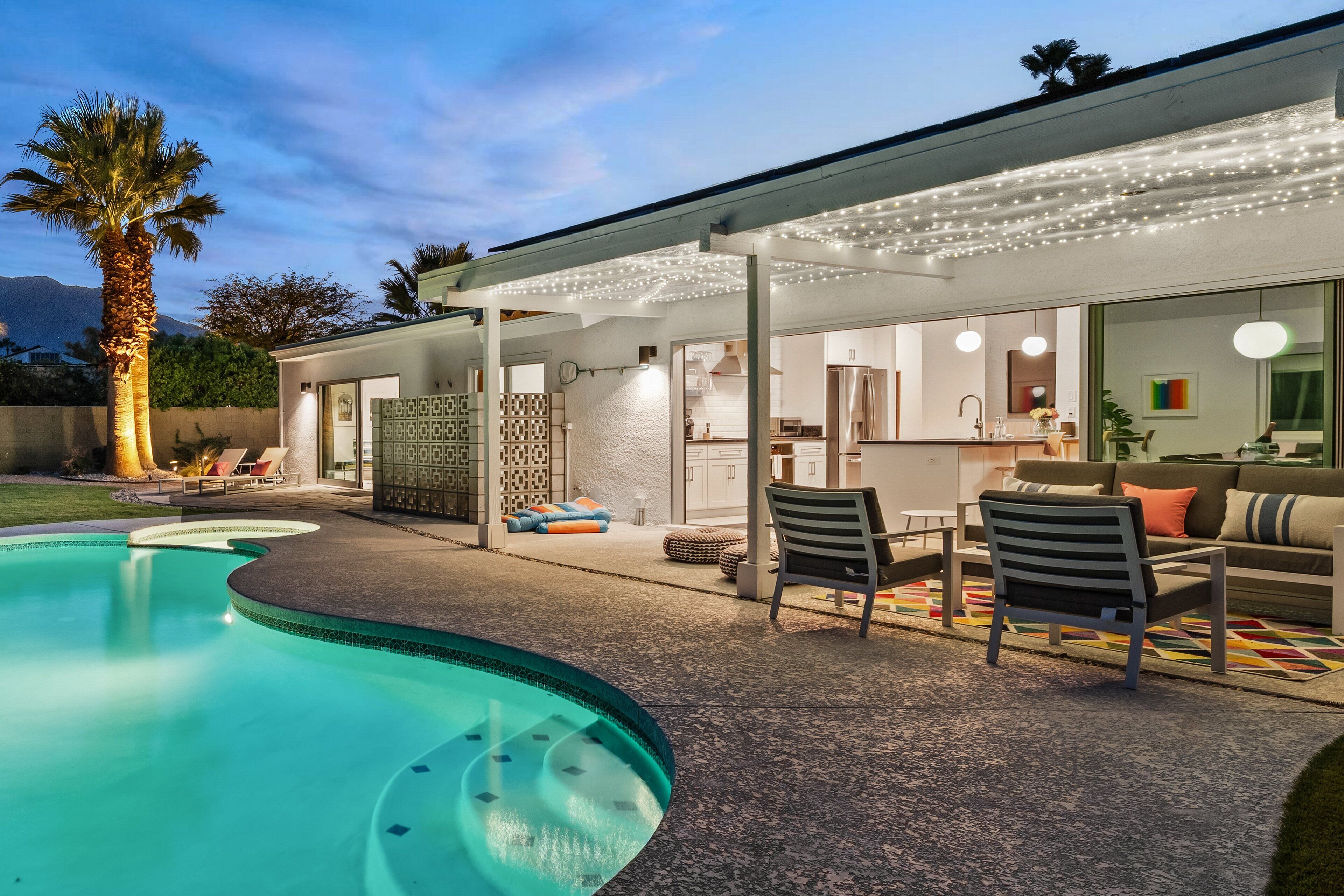 Uma área de estar ao ar livre moderna com piscina, um alpendre coberto e uma cozinha visível através das portas de vidro.