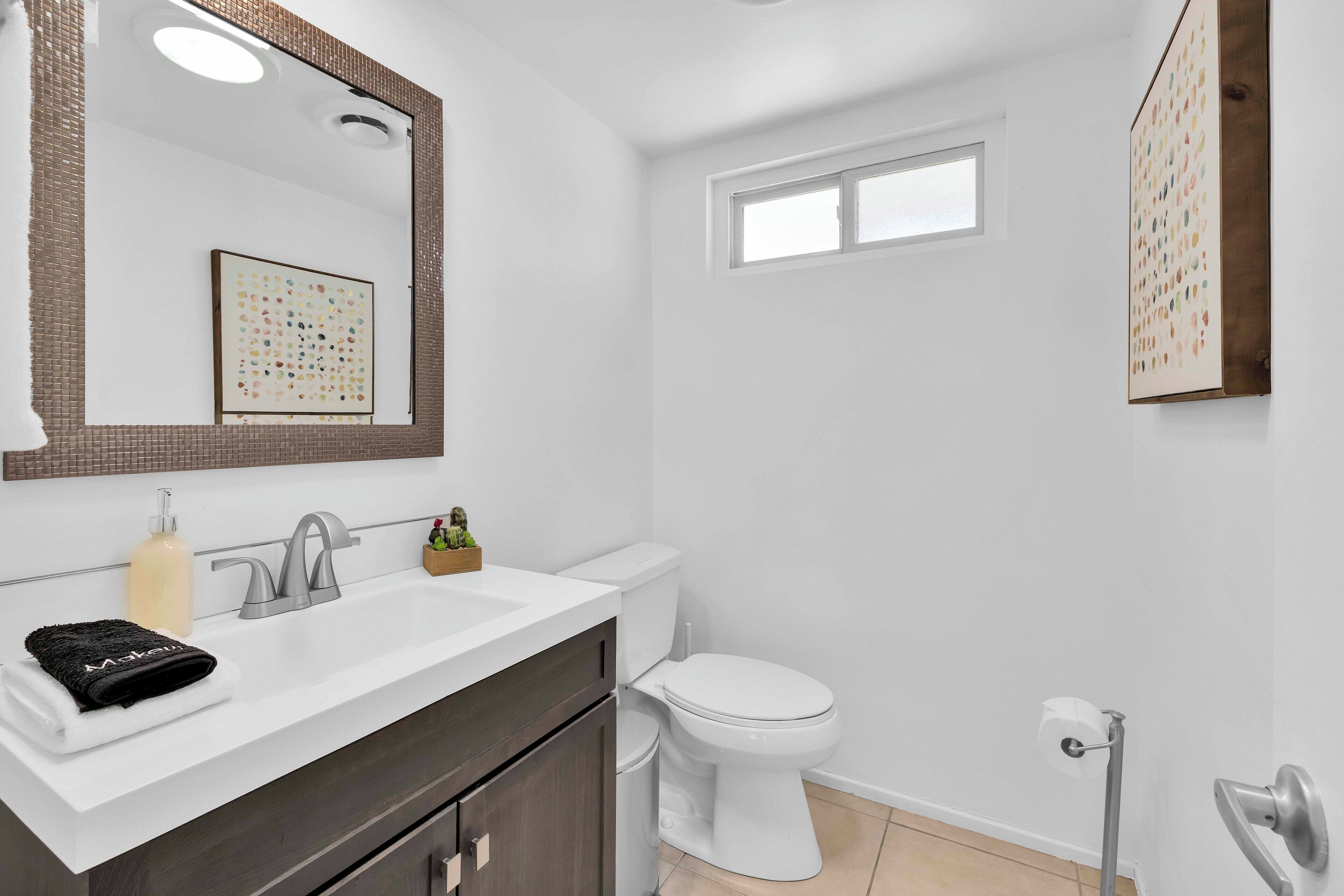 Un baño moderno con un lavamanos blanco, tocador de madera oscura, inodoro blanco y un ventanal pequeño.