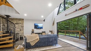 A modern living room with a blue sofa, a stone accent wall, a TV, and a large window overlooking a forest. - Wanderlust A Frame - Lake/ Beach/ Golf/ Pool/ Trails in Alpine Lake (Terra Alta)
