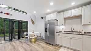 A modern kitchen with white cabinets, a stainless steel refrigerator, and a dining area with a glass door leading to a garden. - Wanderlust A Frame - Lake/ Beach/ Golf/ Pool/ Trails in Alpine Lake (Terra Alta)