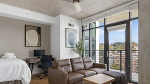 Une chambre d’hôtel moderne équipée d’un lit, d’un canapé en cuir, d’un bureau avec un ordinateur et de grandes fenêtres donnant sur la ville.