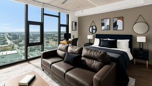 A modern hotel room with a large bed, a leather sofa, and a city view through floor-to-ceiling windows.