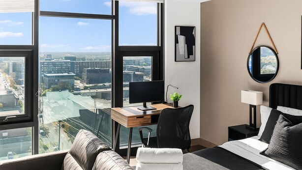 A modern hotel room with a large window offering a city view, a desk with a computer, a comfortable sofa, and a bed with pillows.